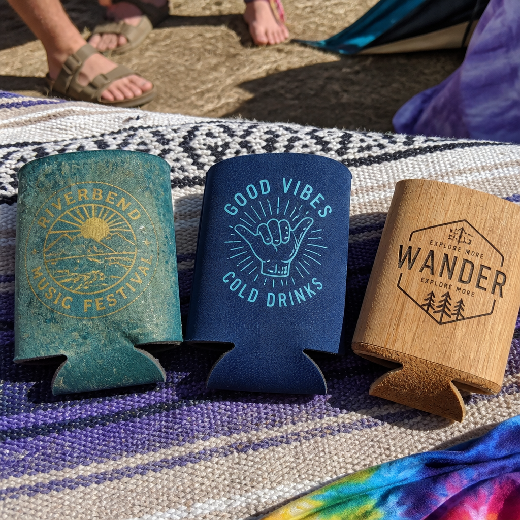 Three festival can sleeves in different materials displayed side-by-side on a blanket at a music festival