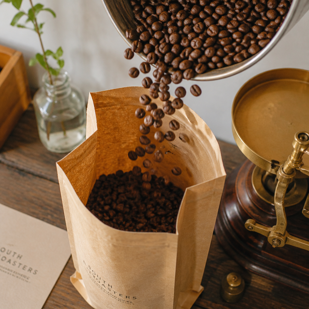 Coffee beans being hand-packed into a kraft paper bag with a brass scale nearby